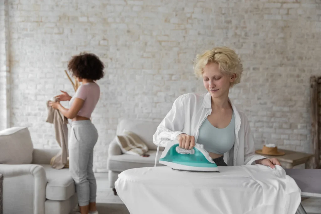 Duas mulheres compartilham tarefas domésticas, passando e dobrando roupas.